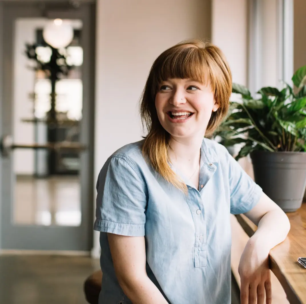 A smiling woman looking at someone else while sitting next to a window ledge.